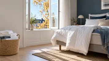Lyst soveværelse med vinduet på vid gab, en hvid dyne lagt til luftning over fodenden af en seng på ben og en kurv med sammenlagt sengetøj